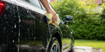 Pessoa lavando um carro preto na garagem de casa, com balde e esponja em um dia ensolarado, demonstrando economia.